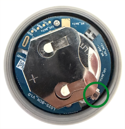 Inside of the L02S-BCN with the case open, showing the circuit board and coin cell battery. The small activation button is circled at the lower right edge of the board.